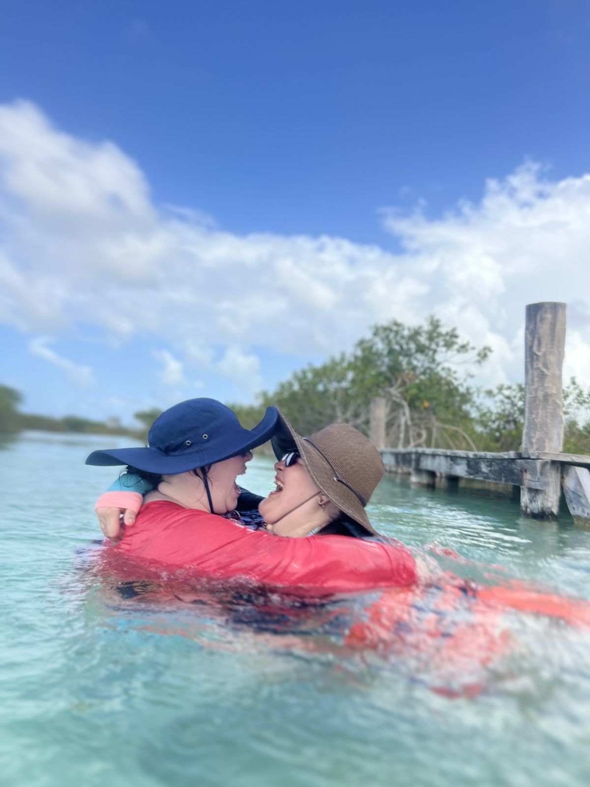 Two queer travelers laughing and embracing while swimming near a dock in Tulum