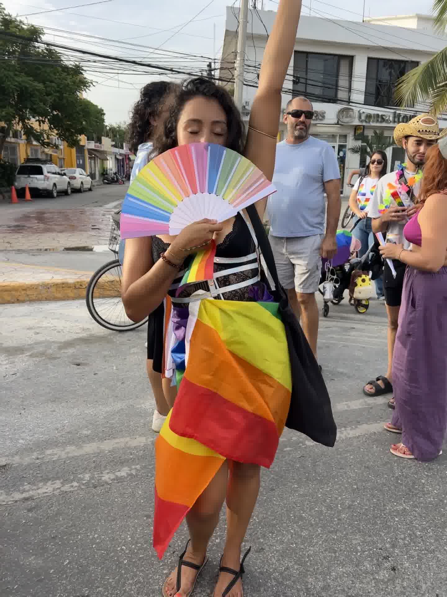 A Pride parade attendee holding a rainbow fan and flag at Tulum Pride